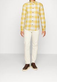 Camisa de cuadros amarillos y blancos de manga larga con botones negros, combinada con pantalones color crema y zapatillas marrones con suela beige.