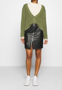 Cardigan fuzzy verde oliva con scollo a V, due bottoni, indossato sopra una maglietta a maniche lunghe color crema, abbinato a una gonna di pelle nera con borchie.