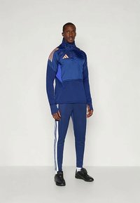 Plavi sportski trenirka izrađena od glatkog materijala; ima zatvarač na kragni, tri pruge niz nogavice i narančaste akcentne Adidas logo. 
