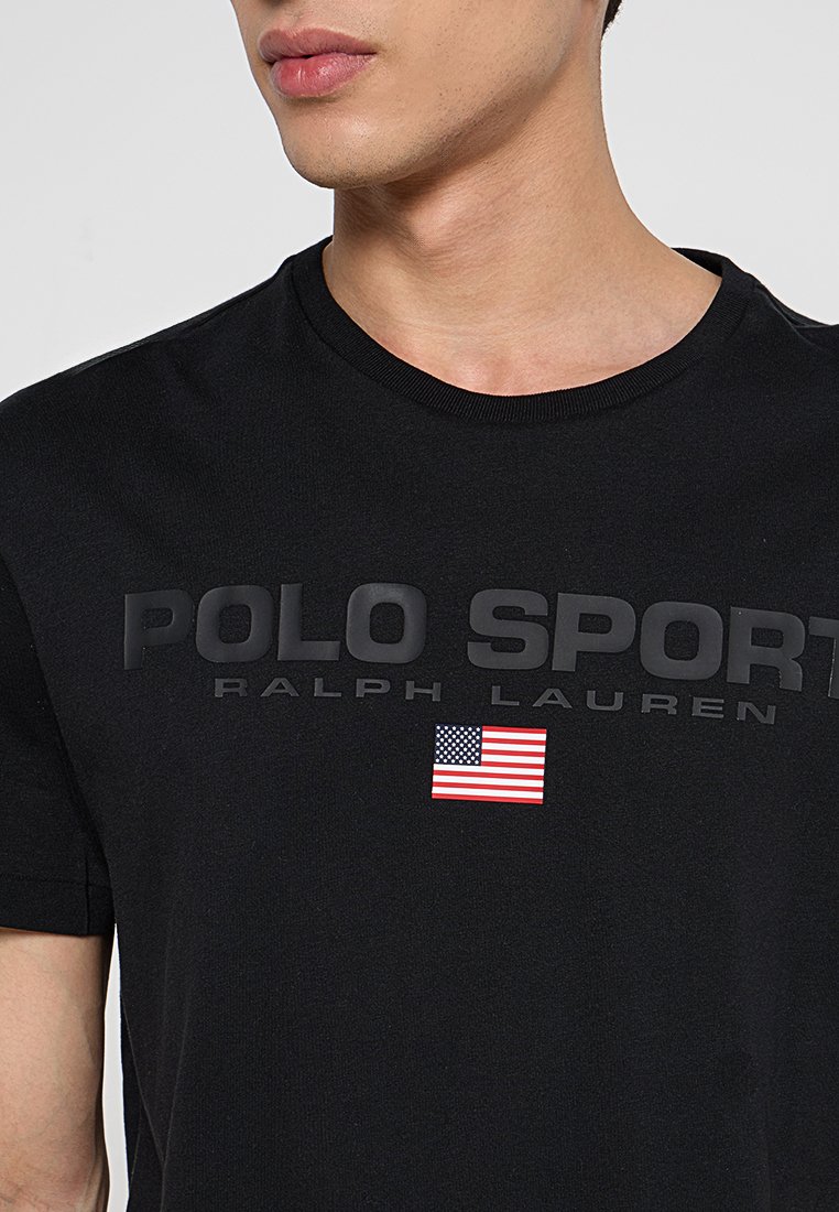Uomo che indossa una maglietta Polo Sport Ralph Lauren nera con logo in rilievo e piccola bandiera americana sul petto.