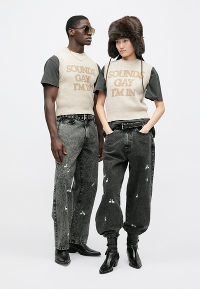 Δύο μοντέλα φορούν casual ρούχα. Ο άντρας έχει ένα μπεζ πλεκτό γιλέκο με κείμενο, γκρι τζιν και γυαλιά ηλίου. Η γυναίκα φοράει τοπ διχρωμίας, χαλαρά γκρι τζιν με μοτίβα λαγού και γούνινο καπέλο.