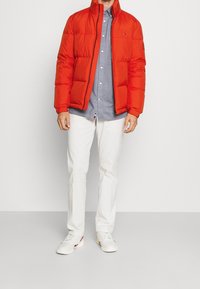 Man i en stark orange puffjacka över en ljusblå skjorta med knappar, med vita byxor och vita sneakers mot en enhetlig bakgrund.