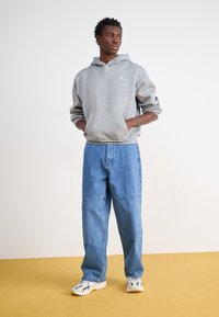 Haut à capuche en coton gris avec poche kangourou et logo, associé à un jean bleu délavé à jambes larges avec détails en panneau. Complété par des baskets sportives.