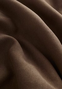 Brązowa tekstura materiału o gładkiej, miękkiej powierzchni. Wyróżniają się delikatnym prążkowaniem i łagodnymi fałdami, podkreślającymi jego elastyczność i opadający kształt.