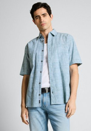 Mężczyzna w jasnoniebieskiej, krótkim rękawie koszuli zapinanej na guziki, na białym podkoszulku, w jasnoniebieskich dżinsach i czarnym pasku, stojący na jednolitym tle.