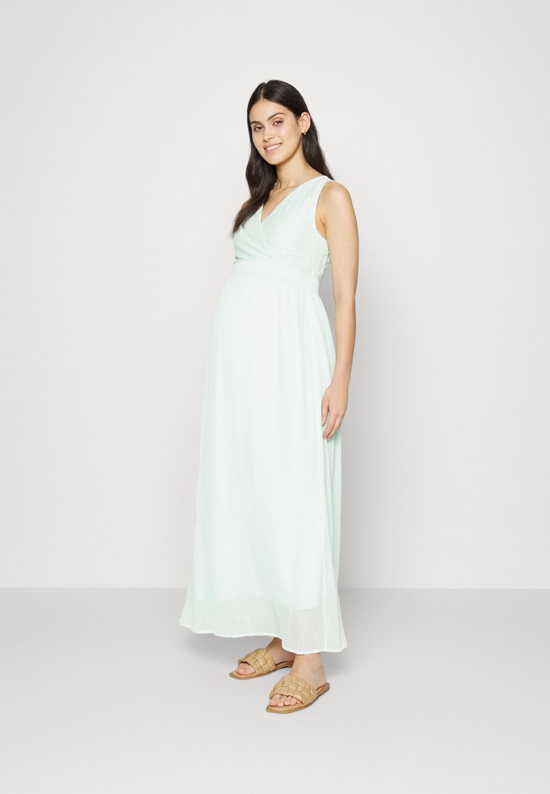 Vestido de maternidade verde menta claro com decote em V e detalhes em renda no corpete, tecido fluido, combinado com sandálias tecidas em tom bege.