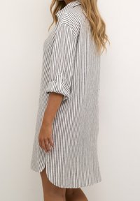 Gestreiftes Hemdkleid in Weiß und Marineblau, mit Kragen, hochgerollten Ärmeln und einer lockeren Passform mit seitlichen Schlitzen für Bewegungsfreiheit.