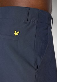 Tamsiai mėlyni šortai su lygiu paviršiumi, turi mažą geltoną erelio logotipą virš kišenės ir šoninės kišenės dizainą.