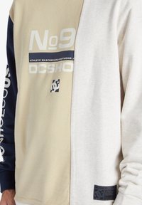 Sudadera con bloques de color que presenta paneles en beige y gris claro, con mangas en navy. En el frontal se exhibe la marca "No.9 DCSHOECOUSA" y un parche con el logo.