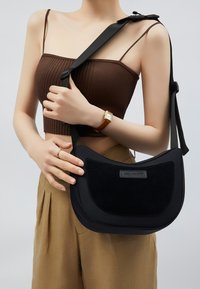 Sort crossbody taske lavet af et glat syntetisk materiale med en suedes frontpanel. Den har en bred skulderrem og en buet form for et moderne design.