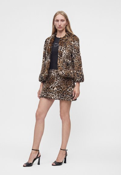 Femme portant une veste en fausse fourrure à imprimé léopard marron et noir avec une jupe assortie, un haut noir et des sandales noires à talons hauts, debout devant un fond blanc.