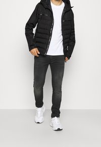 Mand iført en sort hættetrøje over en hvid t-shirt med mørke jeans og hvide sneakers, står foran en ensfarvet lys baggrund.