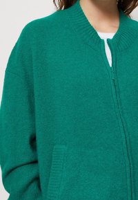 Teal zip-up cardigan i mjukt stickat material, med ribbat krage och manschetter samt sidofickor. Slät struktur med subtil lyster.