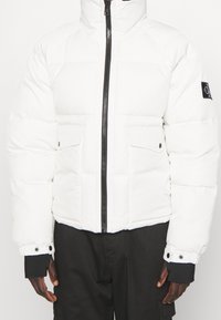 Giacca a vento bianca con zip nera, tasche a soffietto e patch con logo nero sulla manica sinistra, indossata con pantaloni neri.