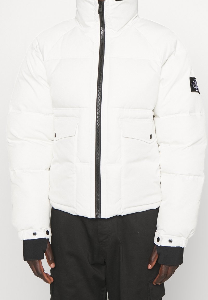 Giacca a vento bianca con zip nera, tasche a soffietto e patch con logo nero sulla manica sinistra, indossata con pantaloni neri.