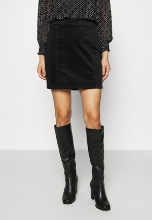 Vrouw draagt een zwarte minirok gecombineerd met kniehoge zwarte leren laarzen en een doorzichtige zwarte blouse met witte stippen.
