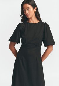 Černé šaty s krátkými nabíranými rukávy, kulatým výstřihem a úzkým pasem. Hladká tkanina s volnou sukní, minimalistický design, bez viditelných vzorů.