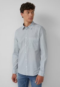 Junger Mann in einem hellblau gemusterten Hemd mit Knöpfen und Jeans, der nach links schaut, vor einem schlichten hellgrauen Hintergrund.
