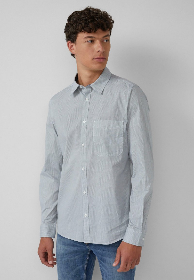 Junger Mann in einem hellblau gemusterten Hemd mit Knöpfen und Jeans, der nach links schaut, vor einem schlichten hellgrauen Hintergrund.