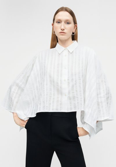 Mladá žena v bielej blúzke na gombíky so širokými, voľne padajúcimi rukávmi a čiernych nohaviciach, stojaca s rukami v vreckách na jednoduchom pozadí.