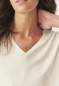 Top cremoso a scollo a V con una consistenza morbida, caratterizzato da una finitura liscia e una vestibilità casual, con dettagli di cucitura a vista vicino al collo.