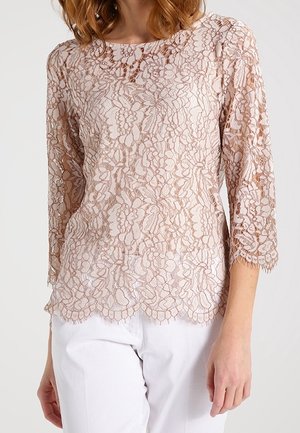 Mujer con una blusa de encaje floral beige de manga larga combinada con pantalones blancos, mostrando bordes delicadamente ondulados en las mangas y el dobladillo.