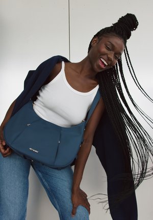 Blauwe schoudertas met een zachte textuur, geometrisch ontwerp en subtiele gouden hardwareaccenten. Gemaakt in combinatie met een witte tanktop en denim jeans.