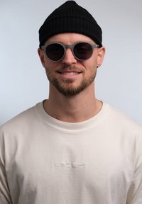 Berretto di maglia nero, occhiali da sole rotondi grigi con lenti scure e una maglietta a maniche lunghe beige chiaro con un sottile logo ricamato sul petto.