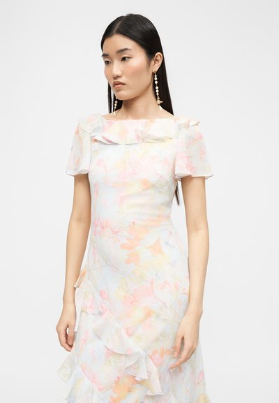 Γυναίκα με μακριά σκούρα μαλλιά που φοράει ένα παστέλ φλοράλ φόρεμα με βολάν και μακριά σκουλαρίκια με μαργαριτάρια, στέκεται μπροστά σε ένα απλό φωτεινό φόντο.