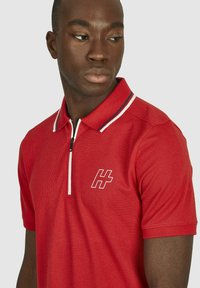 Rode polo-shirt met een ritskraag. Horizontale streepaccenten op de kraag. Heeft een wit logo op de linkerborst. Gemêleerde stof.