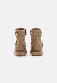 Botines de ante beige con cremallera trasera, que presentan un tacón de madera y correas laterales como detalle. Textura suave y diseño de punta redondeada.
