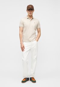 Άνδρας που φοράει μπεζ πόλο πουκάμισο, λευκό παντελόνι, πολύχρωμα παπούτσια και καφέ καπέλο, στέκεται με το ένα χέρι στην τσέπη μπροστά σε απλό φόντο.