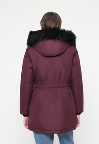 Parka burgund cu glugă din blană sintetică neagră, talie elastică și mâneci lungi, având o textură netedă și un design de lungime medie.