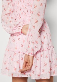 Vestido floral rosa con mangas largas transparentes, bodice fruncido, dobladillo con volantes y una tela ligera y texturizada con pequeños patrones de flores.