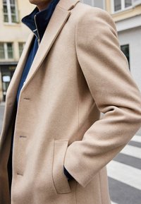 Beige uld-blandet frakke med en skræddersyet pasform, der har en slå-krave, enkelt knaplukning og sidelommer. Lagt over en mørk skjorte.