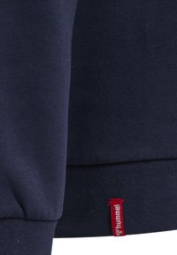El sudadera azul marino presenta un tejido texturizado, puños y dobladillo acanalados, con una pequeña etiqueta de marca roja cerca de la costura inferior.