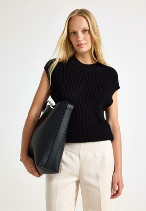 Donna bionda che indossa un top nero senza maniche in maglia e pantaloni beige chiaro tiene una grande borsa tote nera in pelle su sfondo bianco.