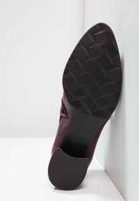 Chaussure en cuir bourgogne avec un bout arrondi, dotée d'une semelle en caoutchouc noir texturé avec des motifs en vagues, affichant la taille "37" sur la semelle.