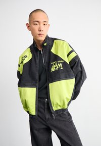 Jaqueta de ganga preta com painéis verde fluorescente, apresentando um colarinho alto, fecho de zíper e logos impressos. Usada com uma blusa preta e jeans.