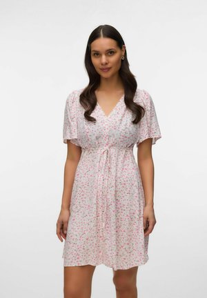 Kvinde med langt mørkt hår iført en hvid knælængde kjole med pink blomsterprint og et bundet bælte i taljen, stående mod en ensfarvet baggrund.