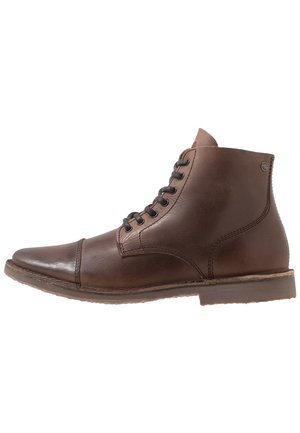 Botas de cuero marrón hasta el tobillo con un acabado suave, punta redonda y ojales de metal. Cuentan con un tacón en contraste color marrón claro y una suela texturizada para mayor agarre.