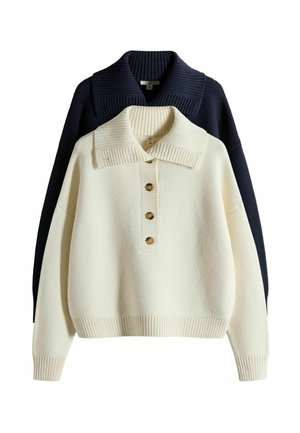 Krém színű kötött pulóver három gombbal és széles gallérral, fekete kötött pulóverre rétegezve, melynek széles bordázott gallérja van.