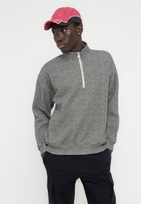 Γκρι πουλόβερ με ψαροκόκκαλο και φερμουάρ στο κολάρο, με μακριά μανίκια και άνετη εφαρμογή· συνδυασμένο με απαλή ροζ καπέλο μπέιζμπολ.