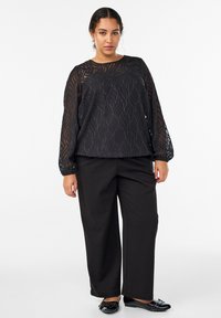 Schwarze durchsichtige Bluse mit strukturiertem Blättermuster, langen Ärmeln und elastischen Manschetten, kombiniert mit weiten schwarzen Hosen und schwarzen flachen Schuhen.