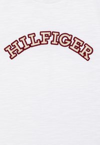 Camiseta blanca con el bordado "HILFIGER" en texto curvado rojo y dorado. Textura suave con un ligero patrón jaspeado. Cuello redondo.