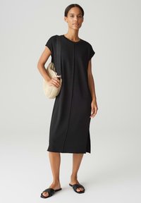 Zwarte midi-jurk met korte mouwen, rechte snit, zijslitsen en een soepele textuur. Gepaard met een geweven tas en platte zwarte sandalen.