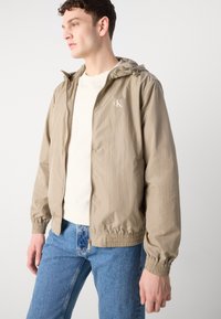 Beige letvægtsjakke med hætte, elastiske manchetter og talje. Har et lille logo på venstre bryst. Bæres over en beige skjorte og blå jeans.