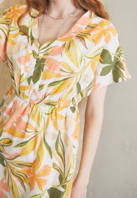 Blommig klänning med V-ringning, korta ärmar och elastisk midja. Tillverkad av lätt tyg med mönster i rosa, gröna och gula blad.