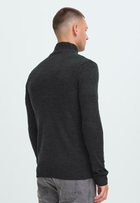 Man die met de rug naar de kamer staat, gekleed in een donkergrijze coltrui en lichtgrijze jeans, tegen een effen lichte achtergrond.