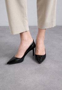 Escarpins en cuir verni noir à talons hauts avec un design à bout pointu et dos ouvert, sur un fond gris clair.